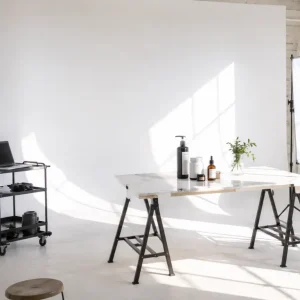 Sunlit photography studio with a white wall, marble work table, assorted bottles, a small plant, and a rolling gear cart on the left
