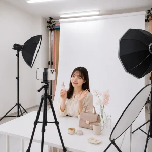 Woman in a photography studio sits at a white table holding a pink perfume bottle, with a camera/phone on a tripod and professional lighting surrounding her; makeup items and a beige handbag on the table as props.