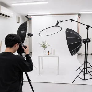Photographer framing a product shoot in a white studio with a table of cosmetics and greenery on a white backdrop