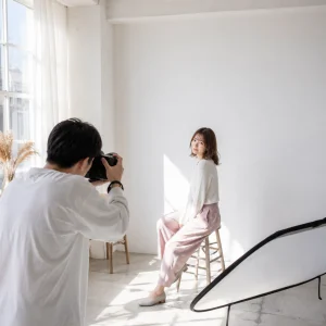 Photographer taking a portrait of a woman seated on a wooden stool in a bright studio with natural light.