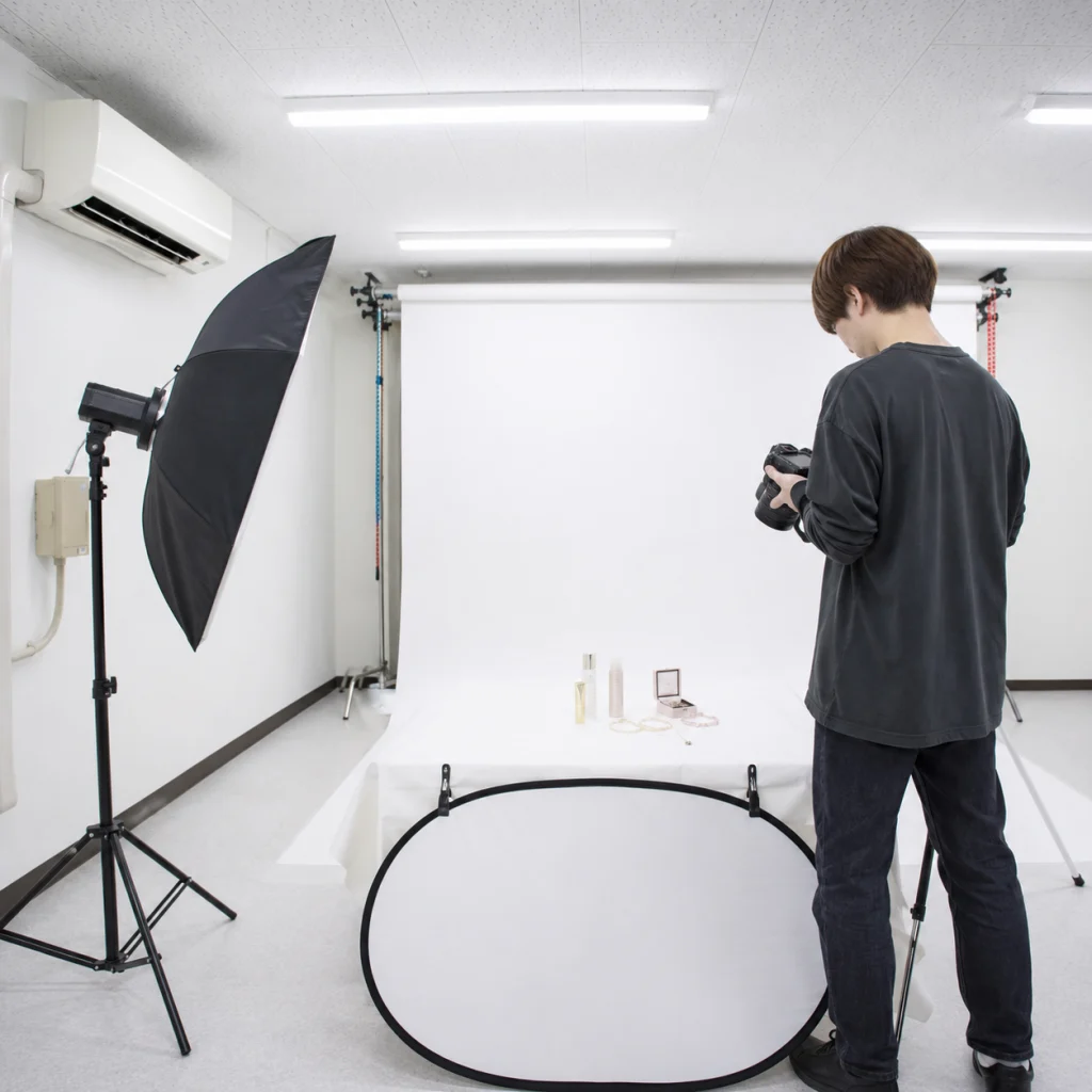 Photographer in studio arranging a camera beside a white backdrop and product display on a table with cosmetics.