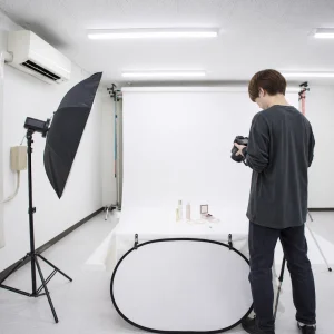 Photographer in studio arranging a camera beside a white backdrop and product display on a table with cosmetics.