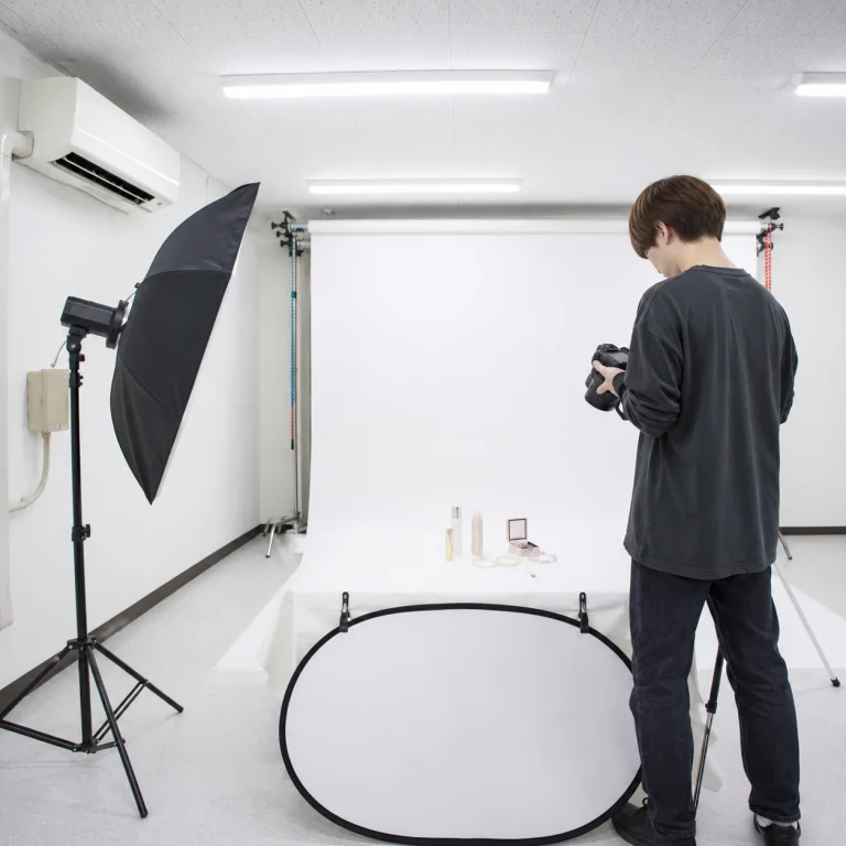 Photographer in studio arranging a camera beside a white backdrop and product display on a table with cosmetics.
