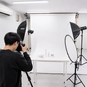 Photographer in a studio shoots skincare products on a white backdrop with bright studio lights and softboxes.