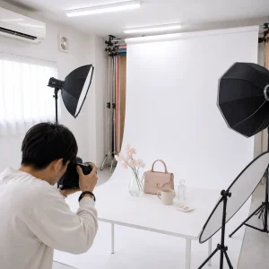Photographer photographs a beige handbag and small items on a white table in a studio with backdrop and lighting gear nearby.