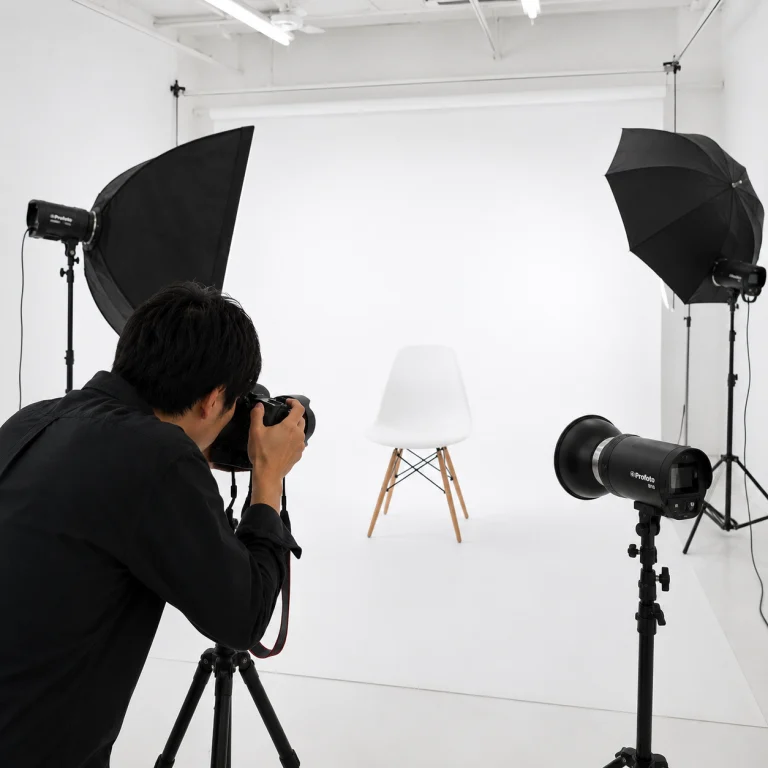 Photographer takes a product shot of a white chair in a bright studio with professional lights and a white backdrop